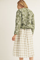 Back of a woman wearing a green floral jacket and plaid skirt on a plain background