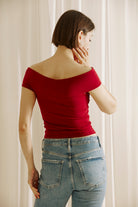 A woman viewed from behind wearing a  burgundy off-shoulder top and blue jeans against a plain background