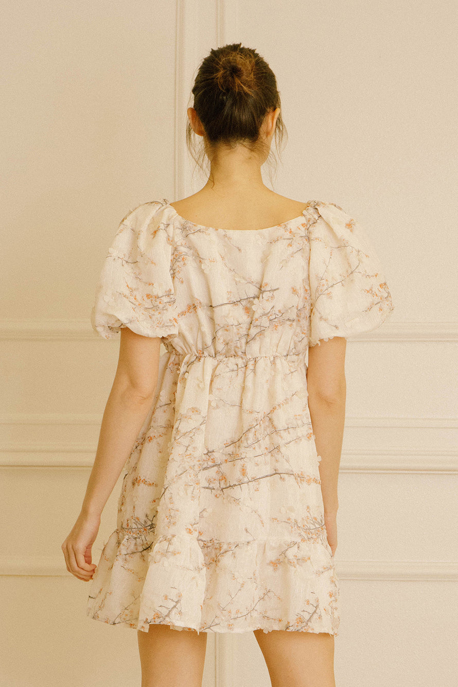 Back view a of woman wearing a cream dress with cherry blossom print against a neutral background