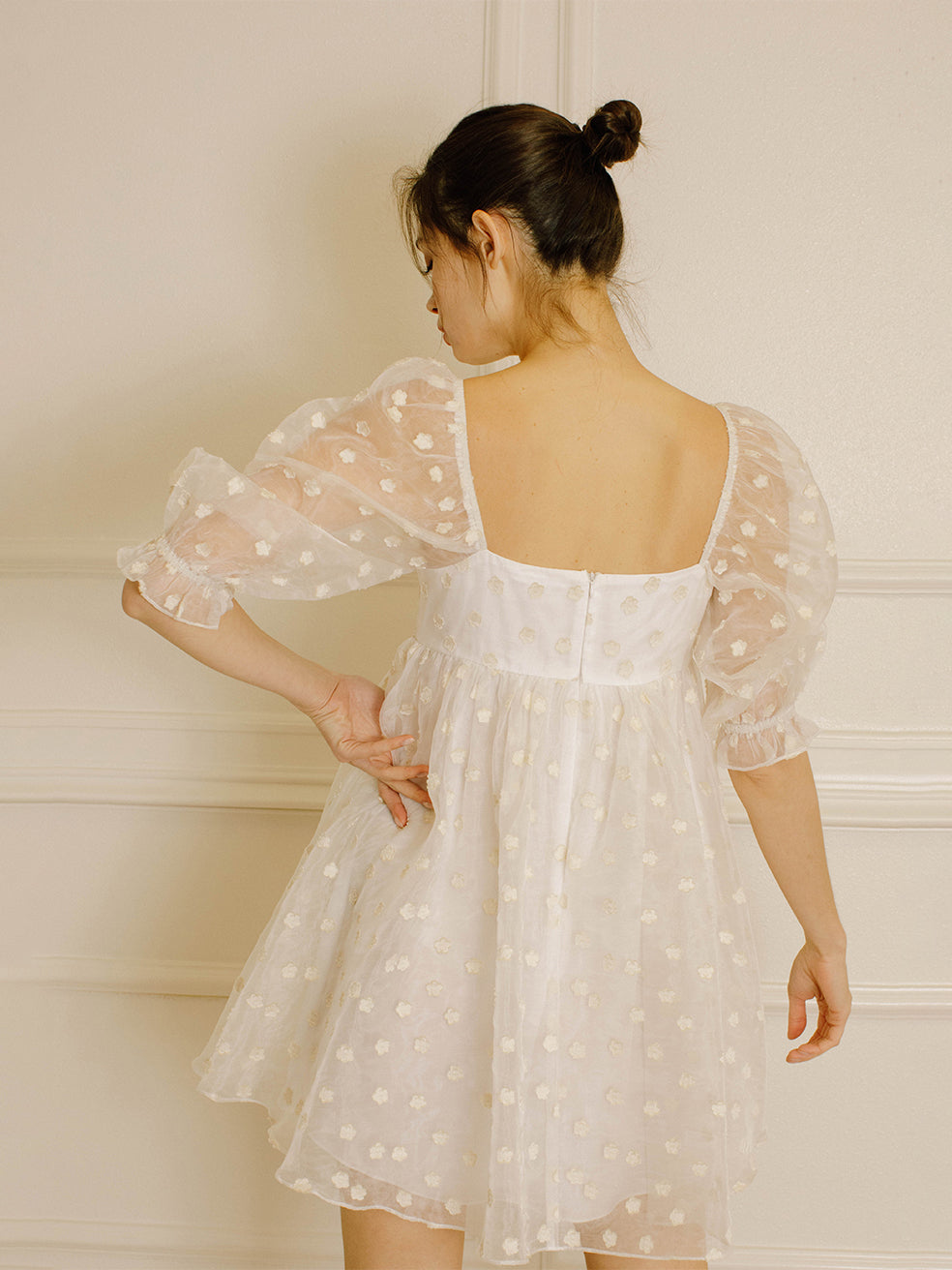 Back view of a woman in a cream polka dot dress against a neutral wall