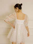 Back view of a woman in a cream polka dot dress against a neutral wall