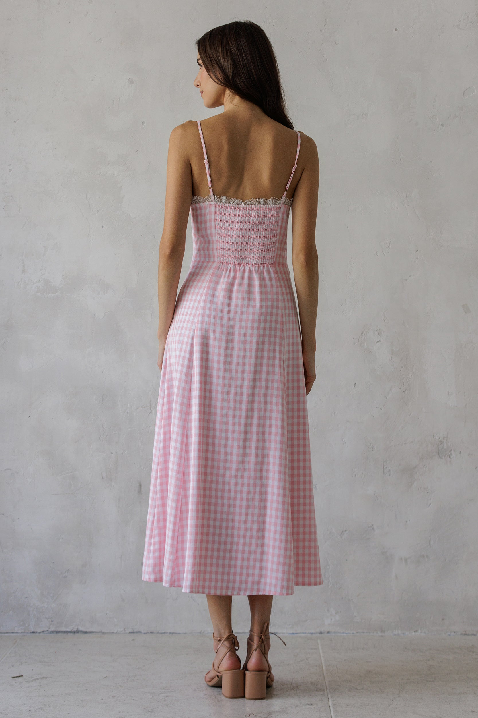 Back of a woman wearing a soft pink gingham pattern midi dress against a grey wall