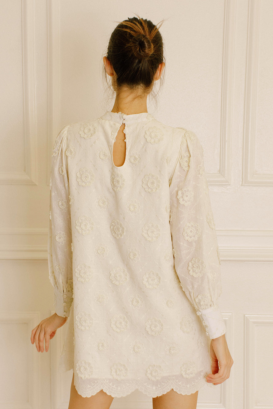 Back view of a woman wearing a cream daisy embroidery mini dress against a neutral wall.