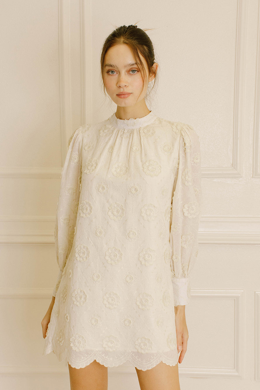 Woman wearing a cream daisy embroidery mini dress against a neutral wall.