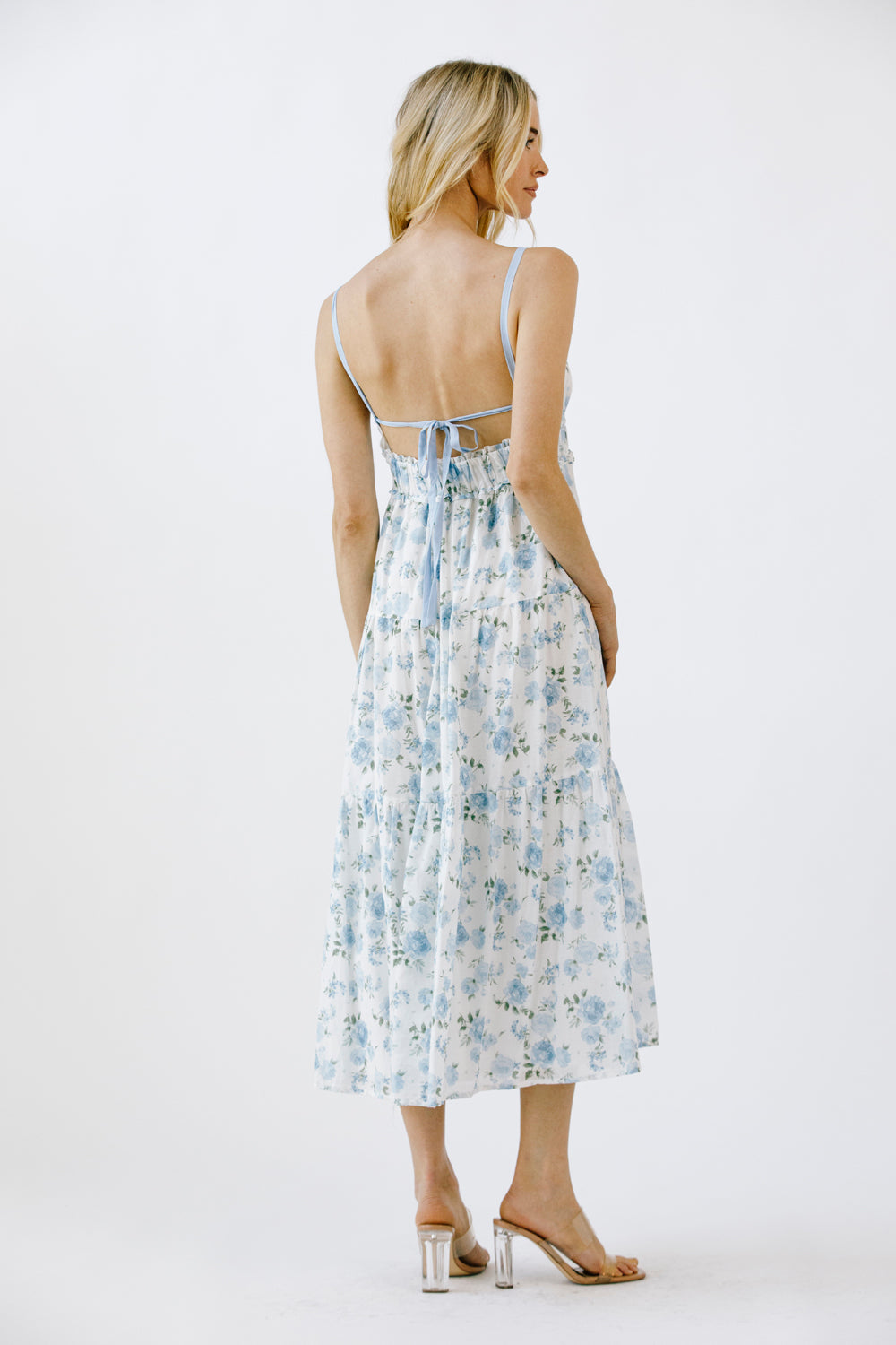 Back view of a woman wearing a blue and white floral midi dress with an open back with self-tie bow on a white background