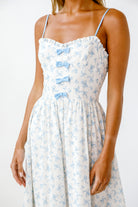 Woman wearing a white dress with blue floral patterns and bow details on a plain background