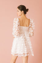 Back view of a woman wearing a white 3D floral mini dress against a pink background