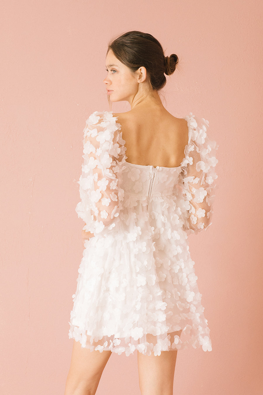 Back view of a woman wearing a white 3D floral mini dress against a pink background