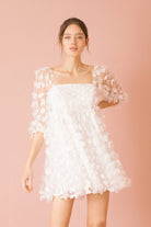 Woman wearing a white 3D floral mini dress against a pink background