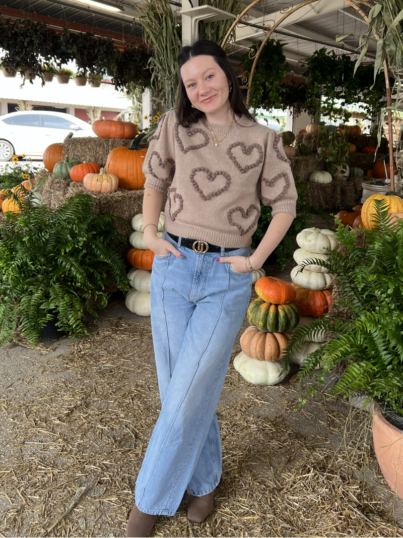 Woman wearing jeans and a brown sweater with hearts standing in a market with pumpkins and plants