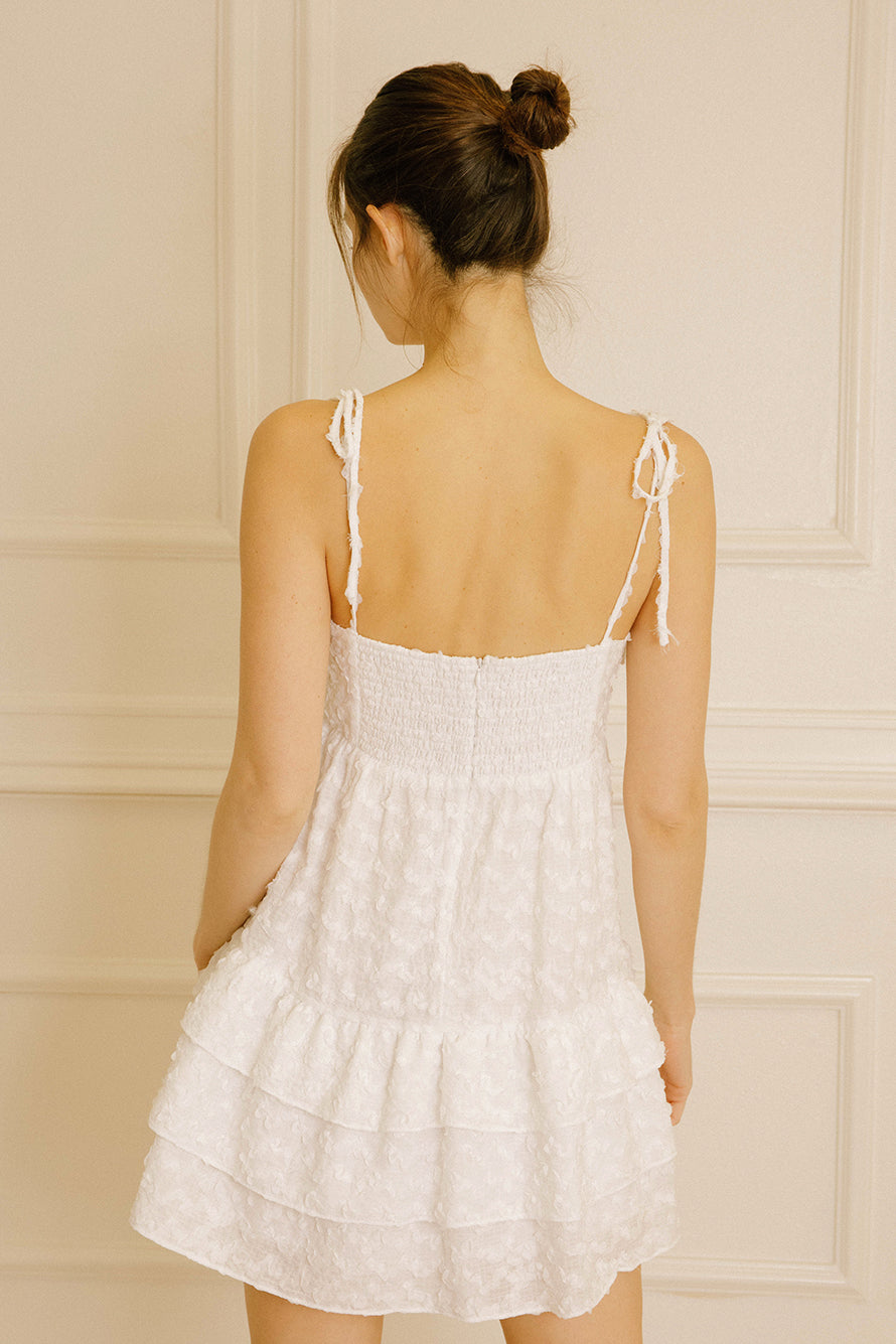 Back view of a woman wearing a white textured dress with adjustable thin straps and back invisible zipper against a beige wall.