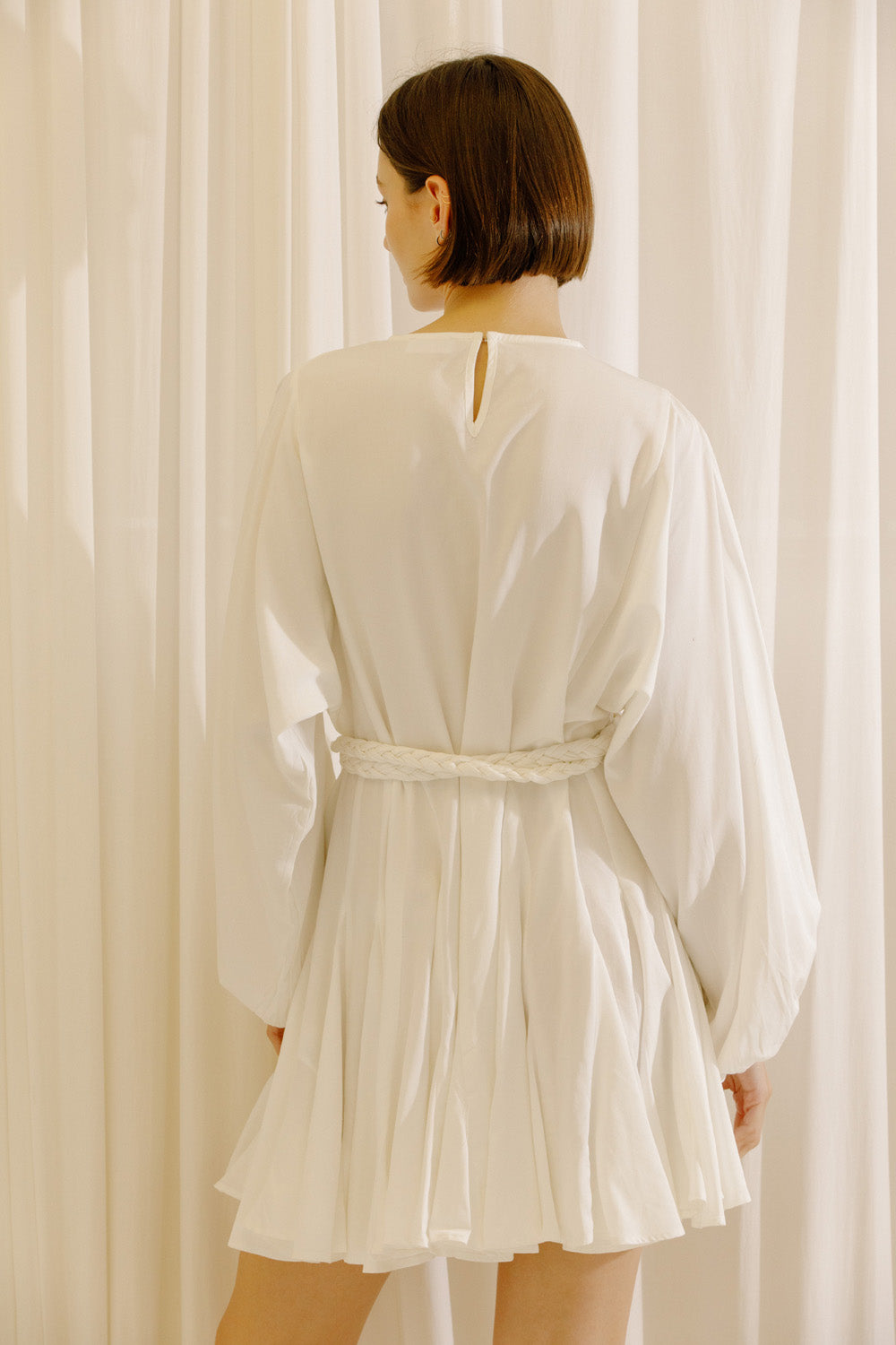 Back view of a woman wearing a white dress with long sleeves and a belt against a beige curtain background