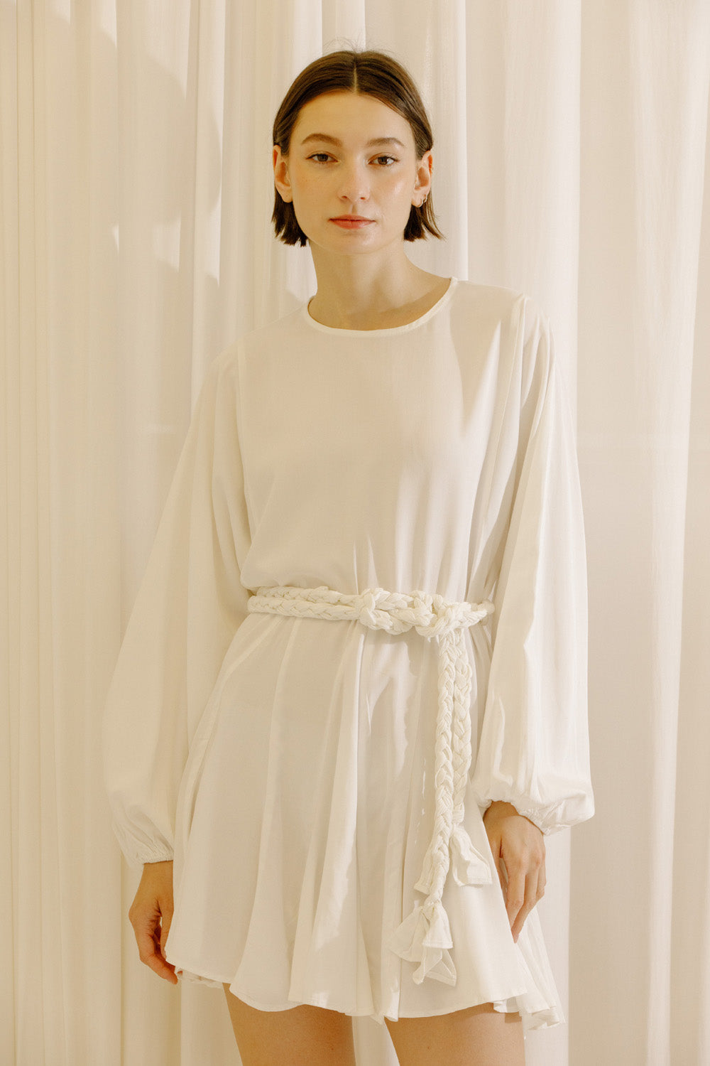 Woman wearing a white dress with long sleeves and a belt against a beige curtain background