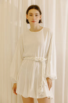 Woman wearing a white dress with long sleeves and a belt against a beige curtain background