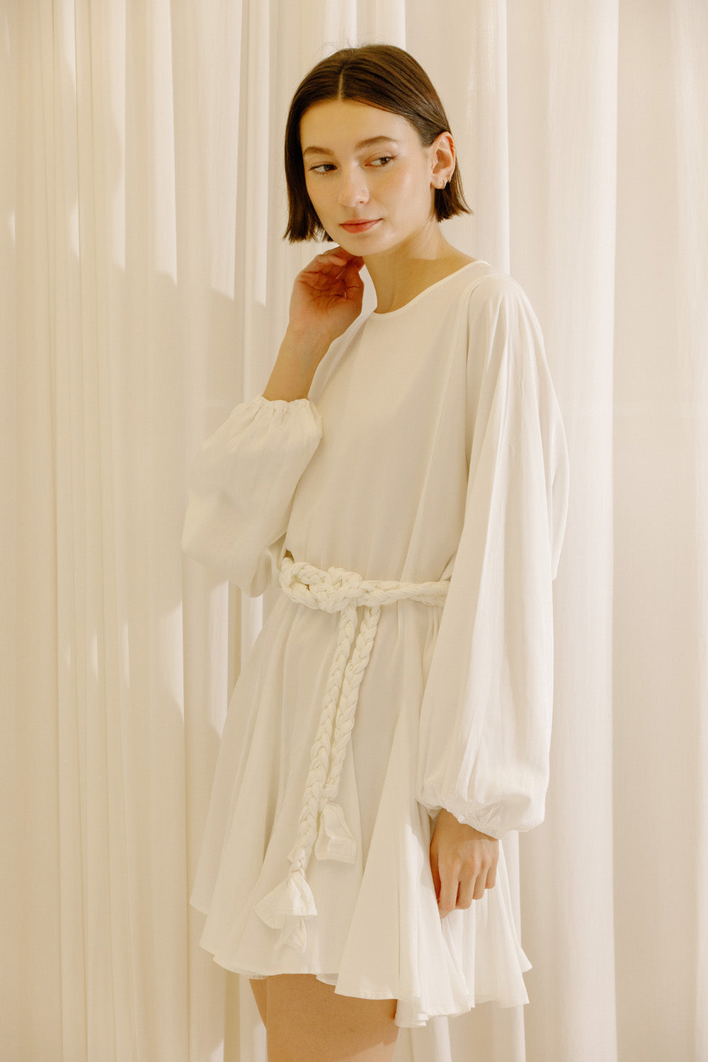 Side view of a woman wearing a white dress with long sleeves and a belt against a beige curtain background