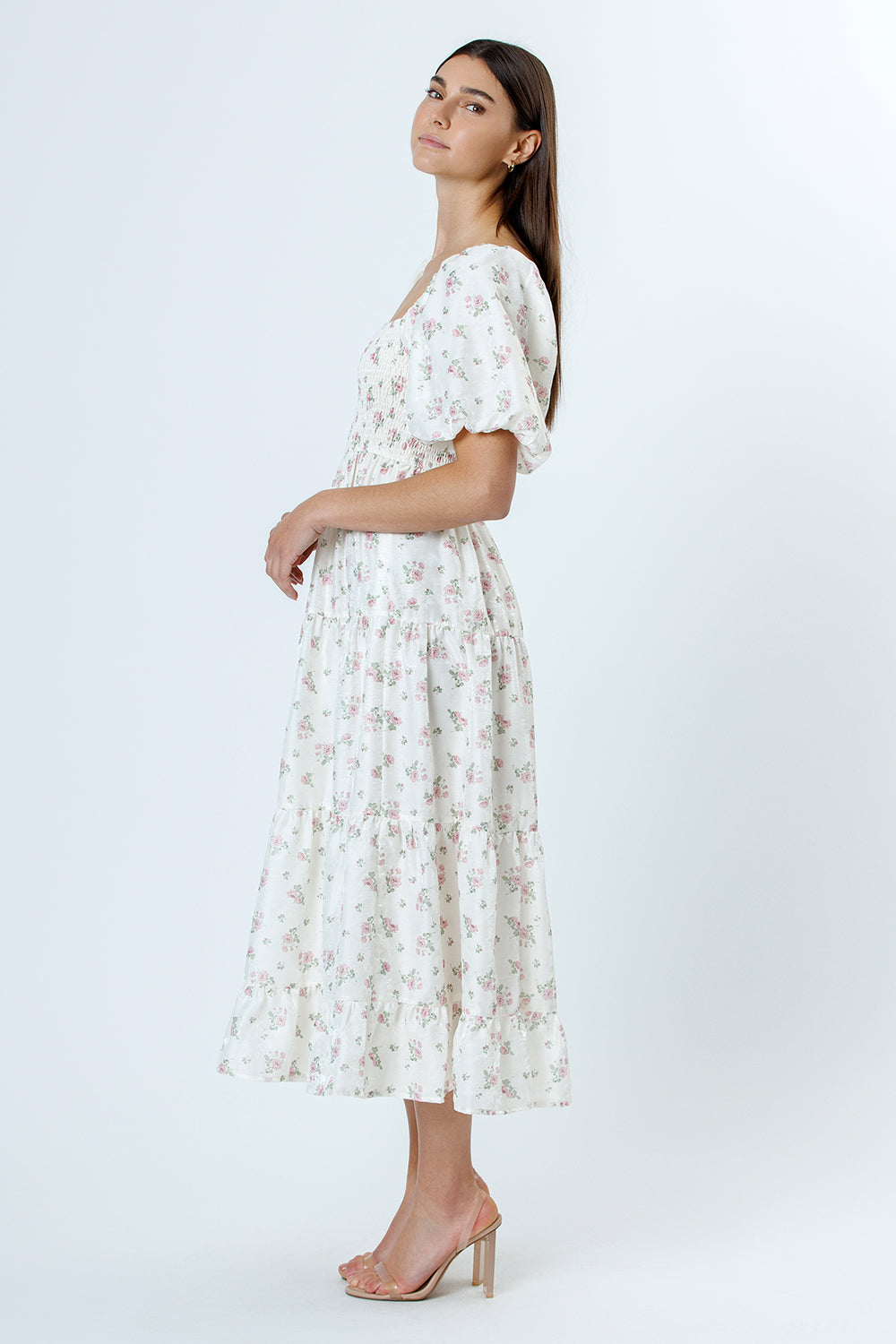 Side view of a woman wearing an ivory with pink rosebuds and flower jacquard print midi dress and beige high-heels on a white background