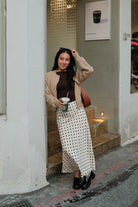 Woman standing outside a building wearing a brown sweater and an ivory and black polka dot skirt, posing for the camera.