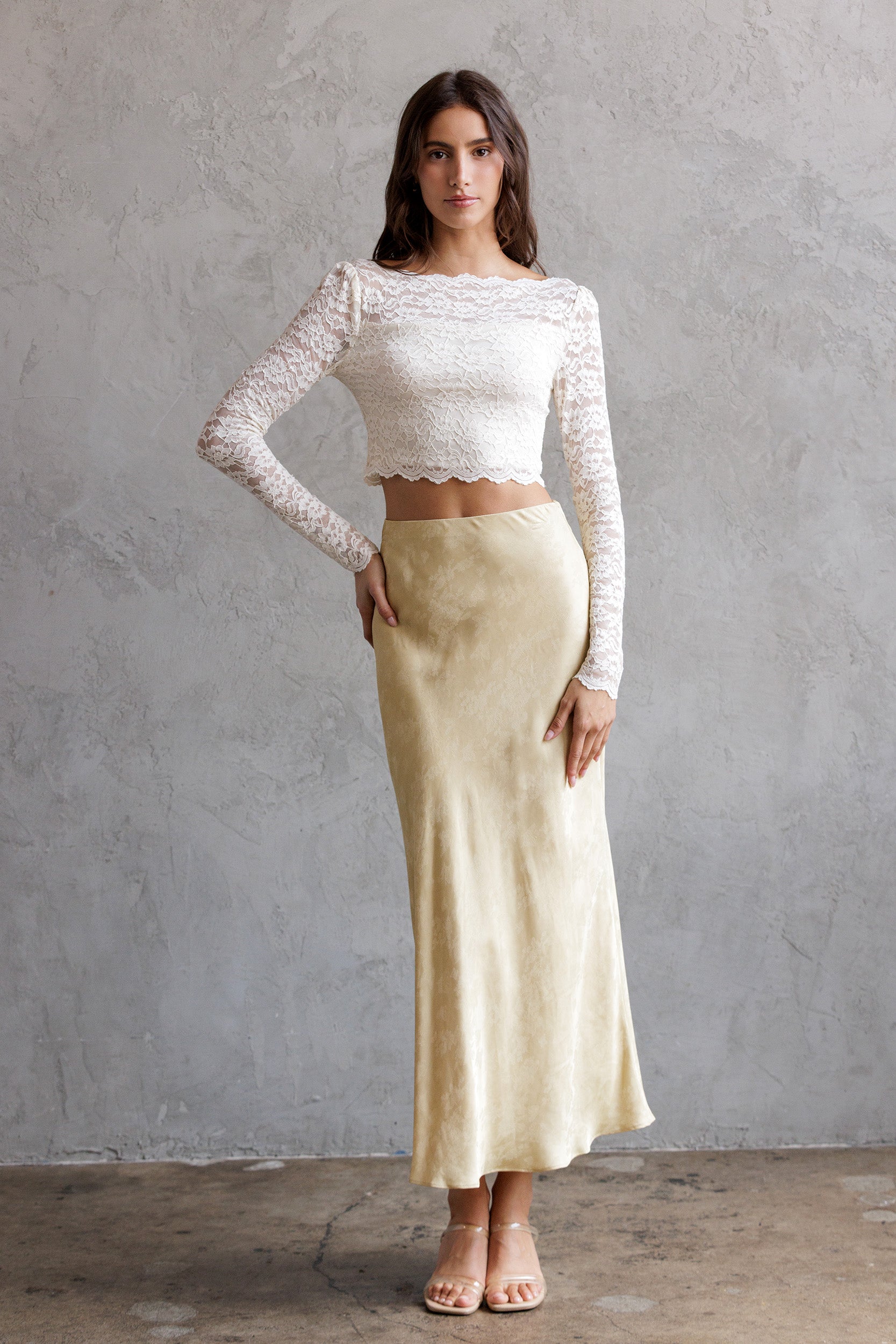 Woman wearing a white lace top and light yellow skirt against a gray wall.