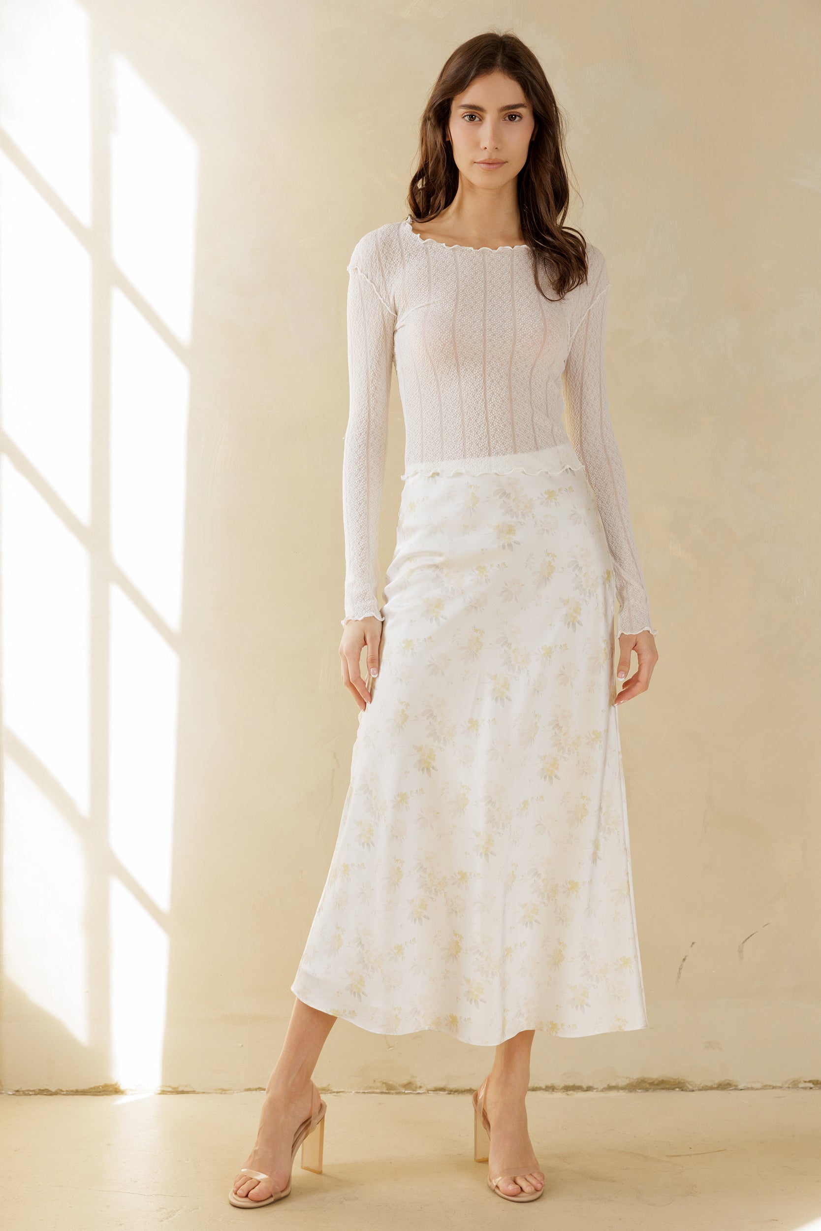 Woman wearing a white long-sleeve top and a white floral skirt in a bright room.