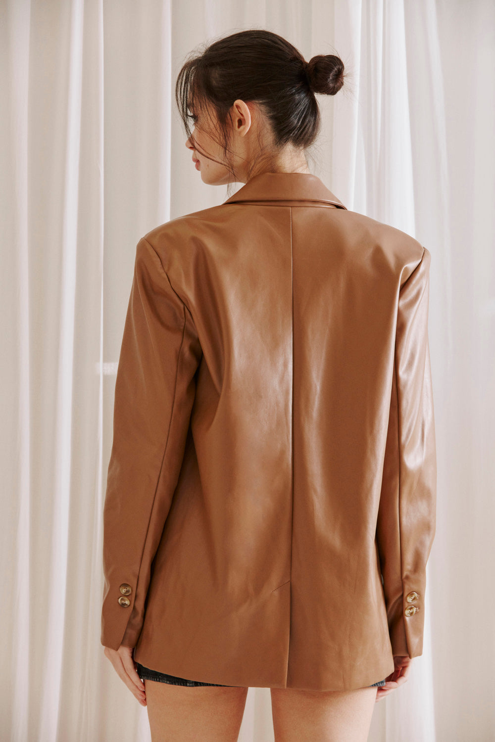 Back view of a woman wearing a brown faux leather blazer over a black dress against a white curtain background