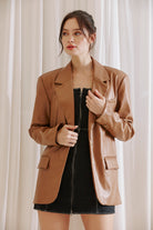 Woman wearing a brown faux leather blazer over a black dress against a white curtain background