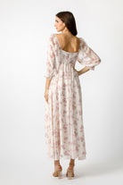 A woman modeling a long-sleeved maxi dress with a pink floral print on a cream background, shown from the back against a light gray studio backdrop.