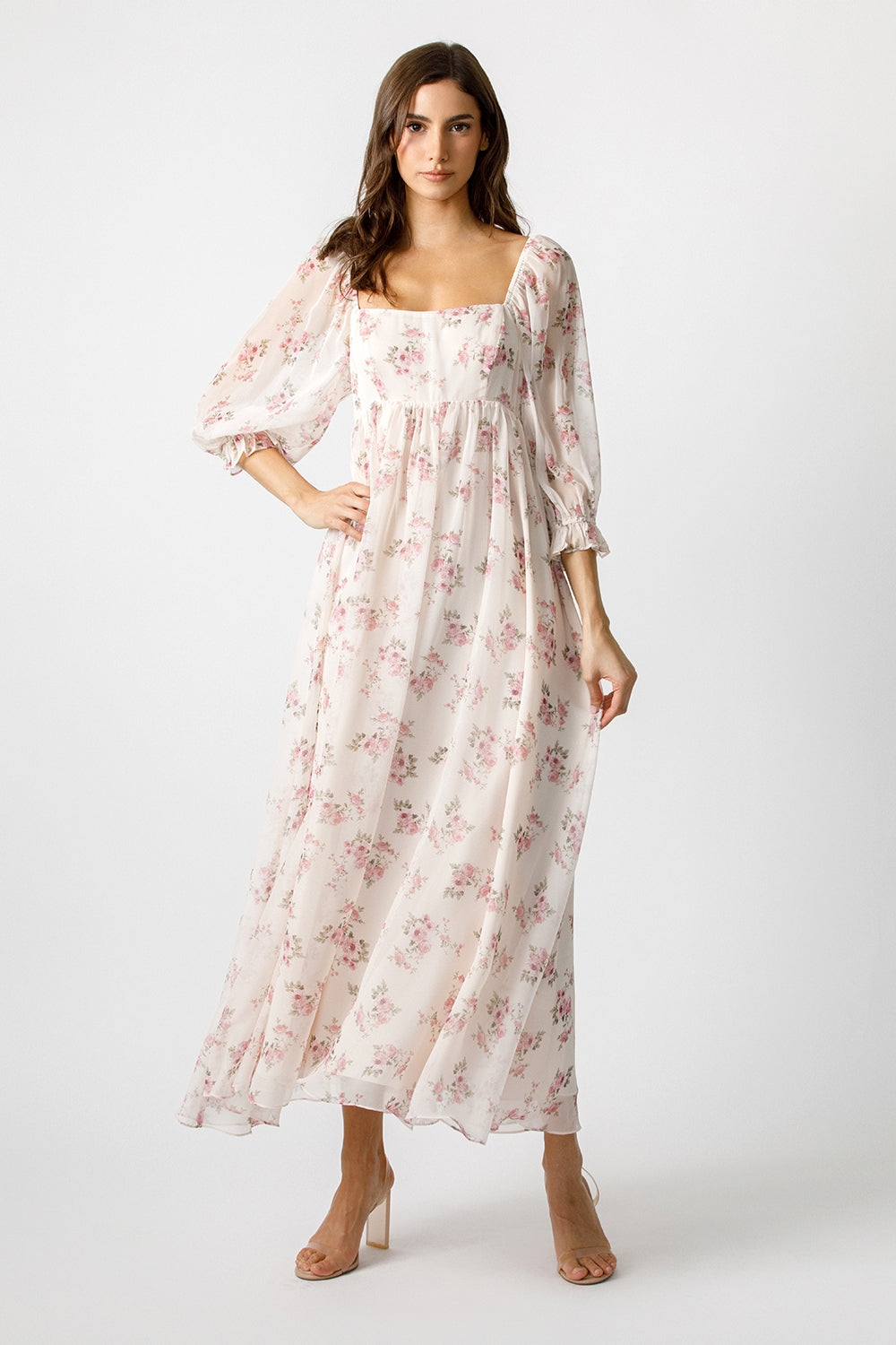 A woman modeling a long, flowy midi dress with a square neckline, puff sleeves, and an allover pink floral print on a light cream background, photographed against a plain light gray studio backdrop.
