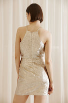 Back of a woman wearing a gold mini dress with an invisible zipper against a beige curtain background