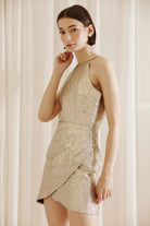Side view of a woman wearing a gold mini dress against a beige curtain background