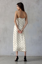 A woman shown from the back wearing an ivory midi slip dress with black polka dots and thin spaghetti straps, paired with black high heels, standing against a textured gray concrete wall.