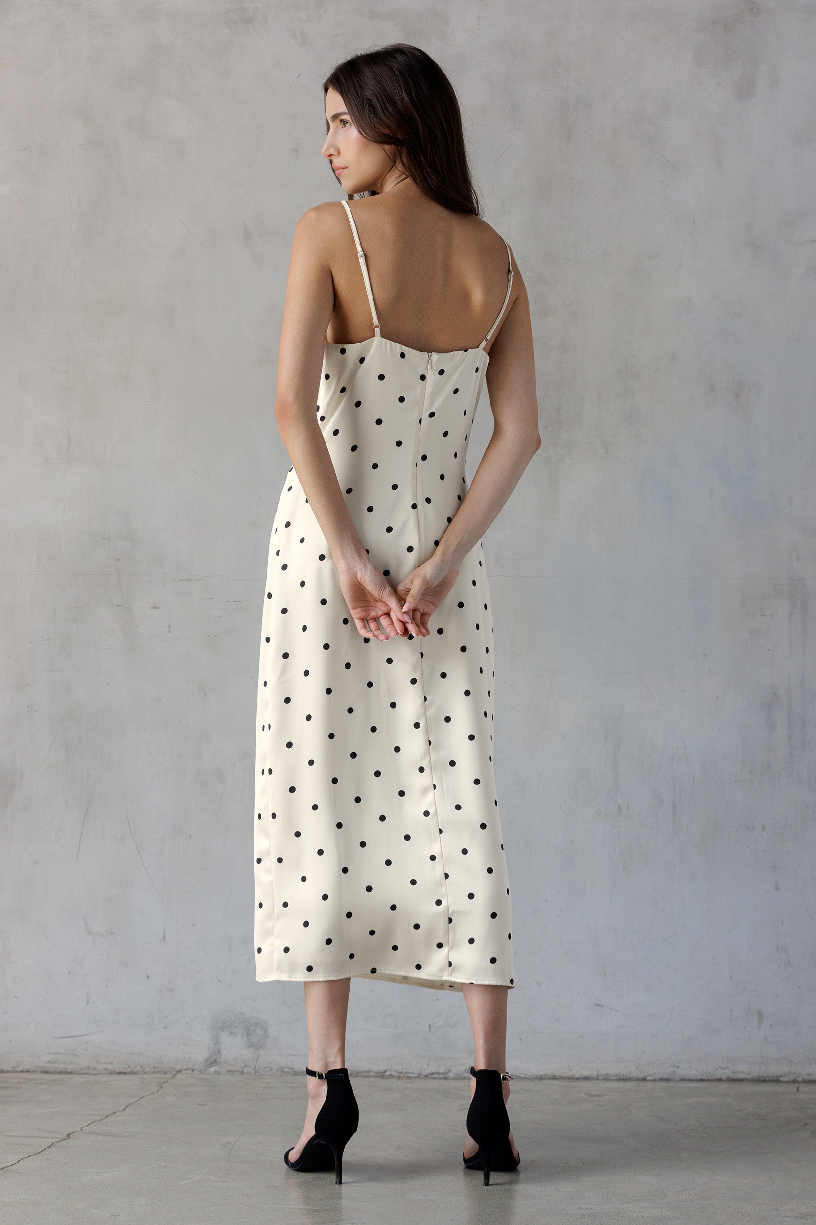 A woman shown from the back wearing an ivory midi slip dress with black polka dots and thin spaghetti straps, paired with black high heels, standing against a textured gray concrete wall.