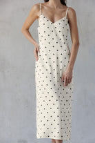 A woman wearing an ivory spaghetti-strap slip dress with a black polka dot pattern, standing against a textured gray wall.
