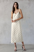 A woman modeling an ivory spaghetti-strap midi dress with black polka dots, paired with black strappy high-heeled sandals, standing against a textured gray concrete wall.