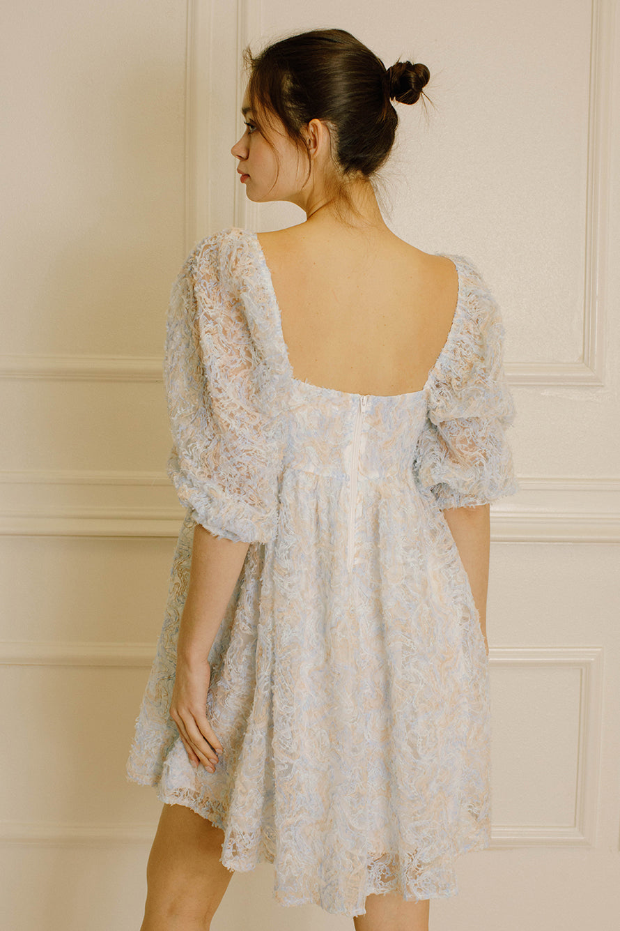 Back view of a woman wearing a blue and cream textured dress against a neutral wall.