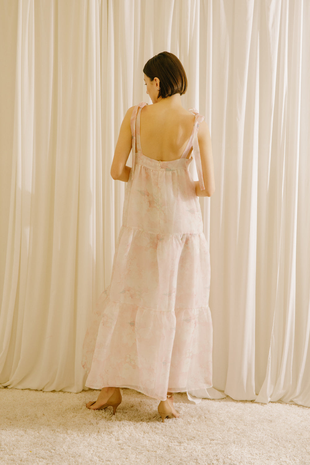 Back view of a woman in a light pink dress standing against a white curtain backdrop