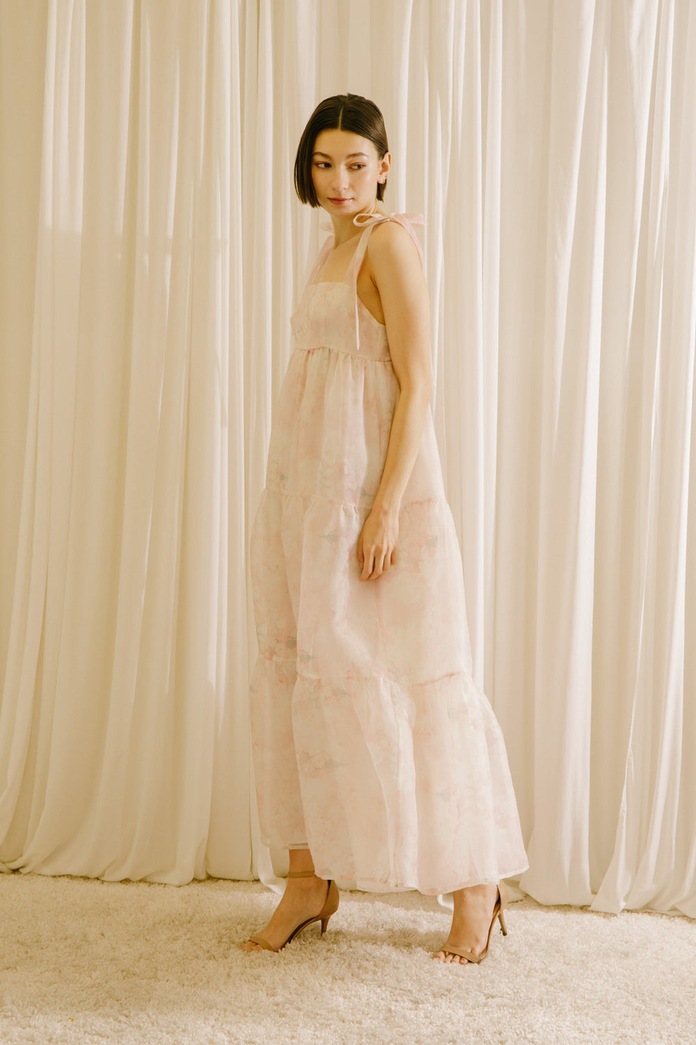 Side view of a woman in a light pink dress standing against a white curtain backdrop
