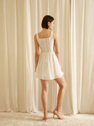 Back view of a woman wearing a white mini dress with rose appliqués and a structured corset bodice against a neutral wall