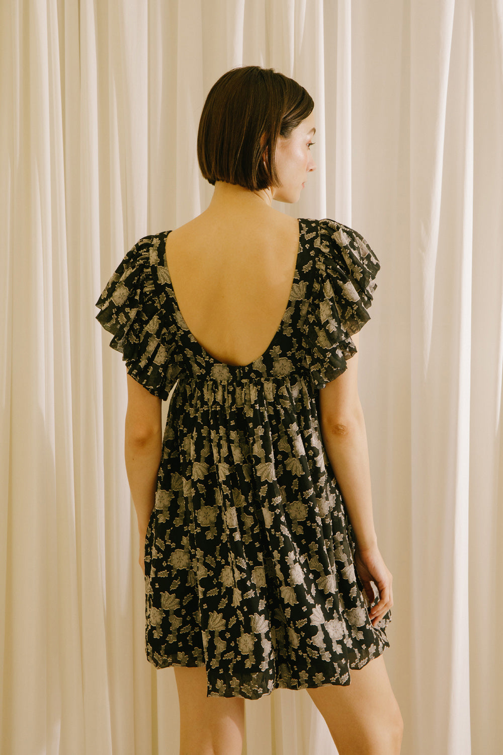 Back view of a woman wearing a black dress with a beige pattern, flutter sleeves and a v-neckline against a beige curtain background