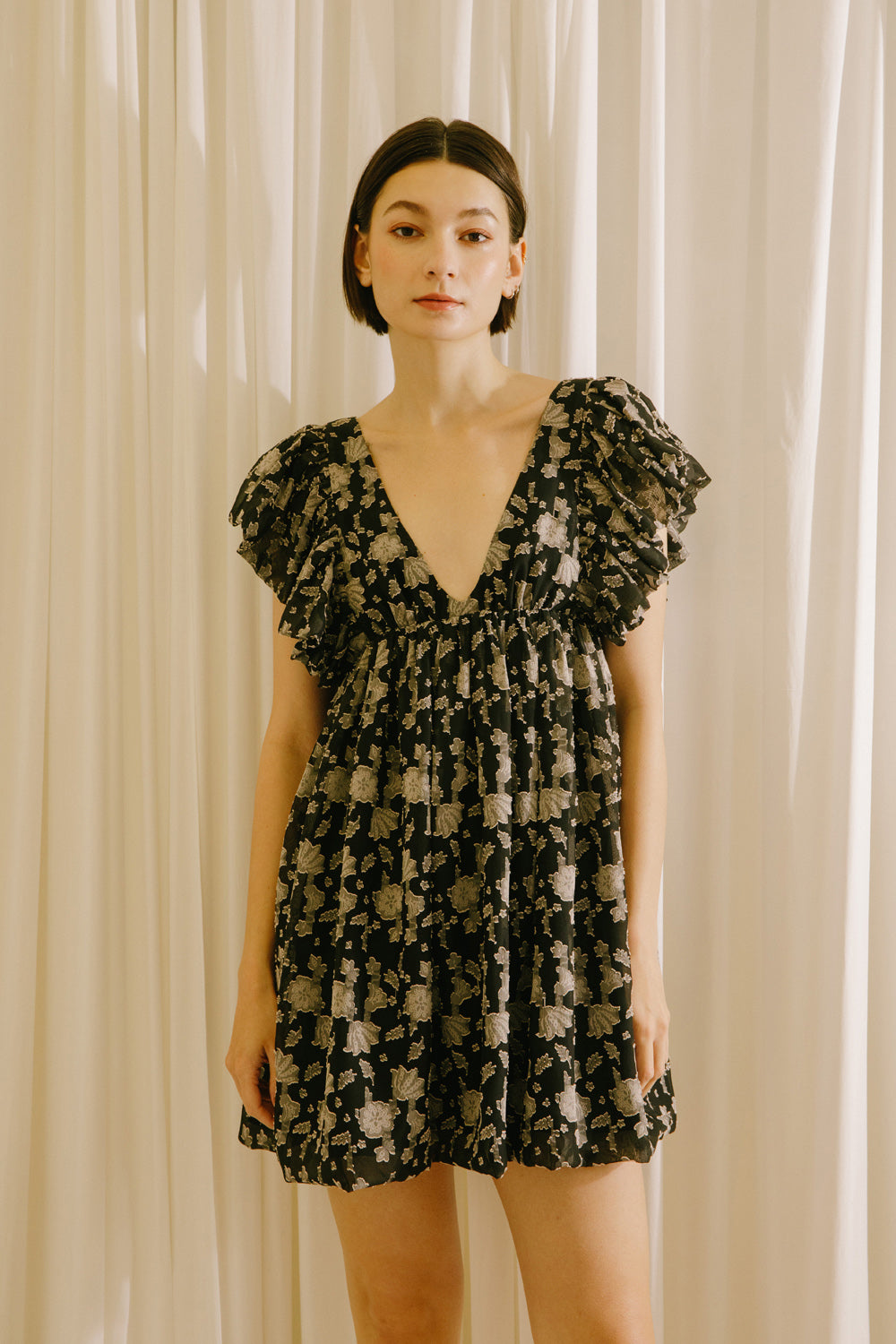 Woman wearing a black dress with a beige pattern, flutter sleeves and a v-neckline against a beige curtain background