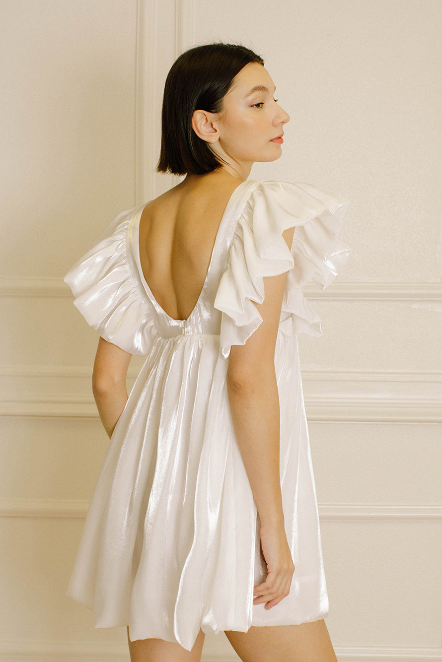 Back view of a woman wearing a white dress with ruffled sleeves against a beige wall.