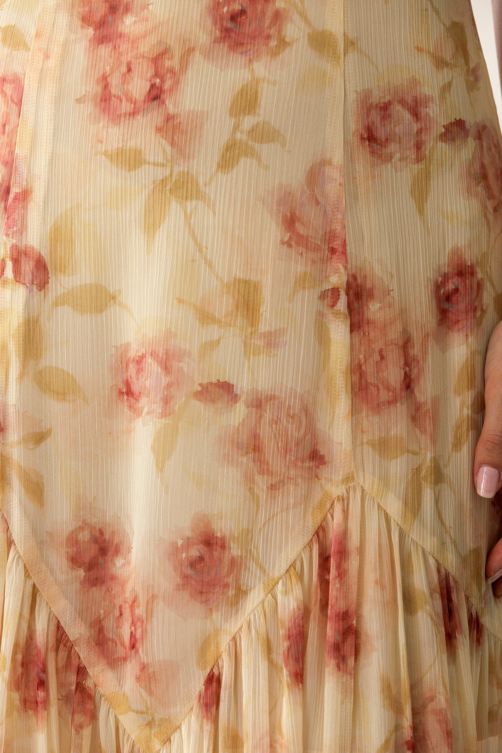 Close-up of a sheer cream-colored fabric with a pink and beige floral print, featuring a ruffled hem and a hand holding the side.