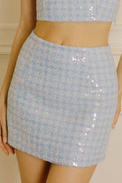 A light blue tweed mini skirt with sequined details, shown on a model. The image is cropped to show the waist, hands, and legs of the model wearing the outfit.