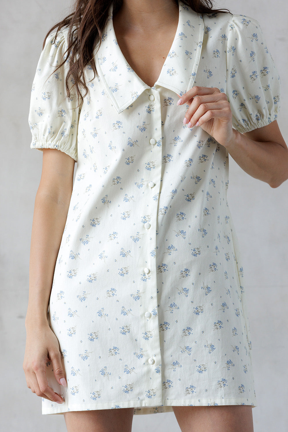 A person wearing a short-sleeve button-front shirt dress with a small blue floral print on a warm off-white background, shown against a light gray wall.