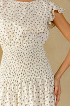 Close-up of a woman wearing a cream-colored smocked dress with a small blue and green floral print. The dress features ruffled cap sleeves and a gathered waist.
