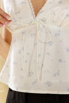 Close-up of an ivory  sleeveless blouse with a light blue floral print, featuring a lace-trimmed V-neckline, gathered bust, and a front tie detail. A hand is shown holding the side fabric.