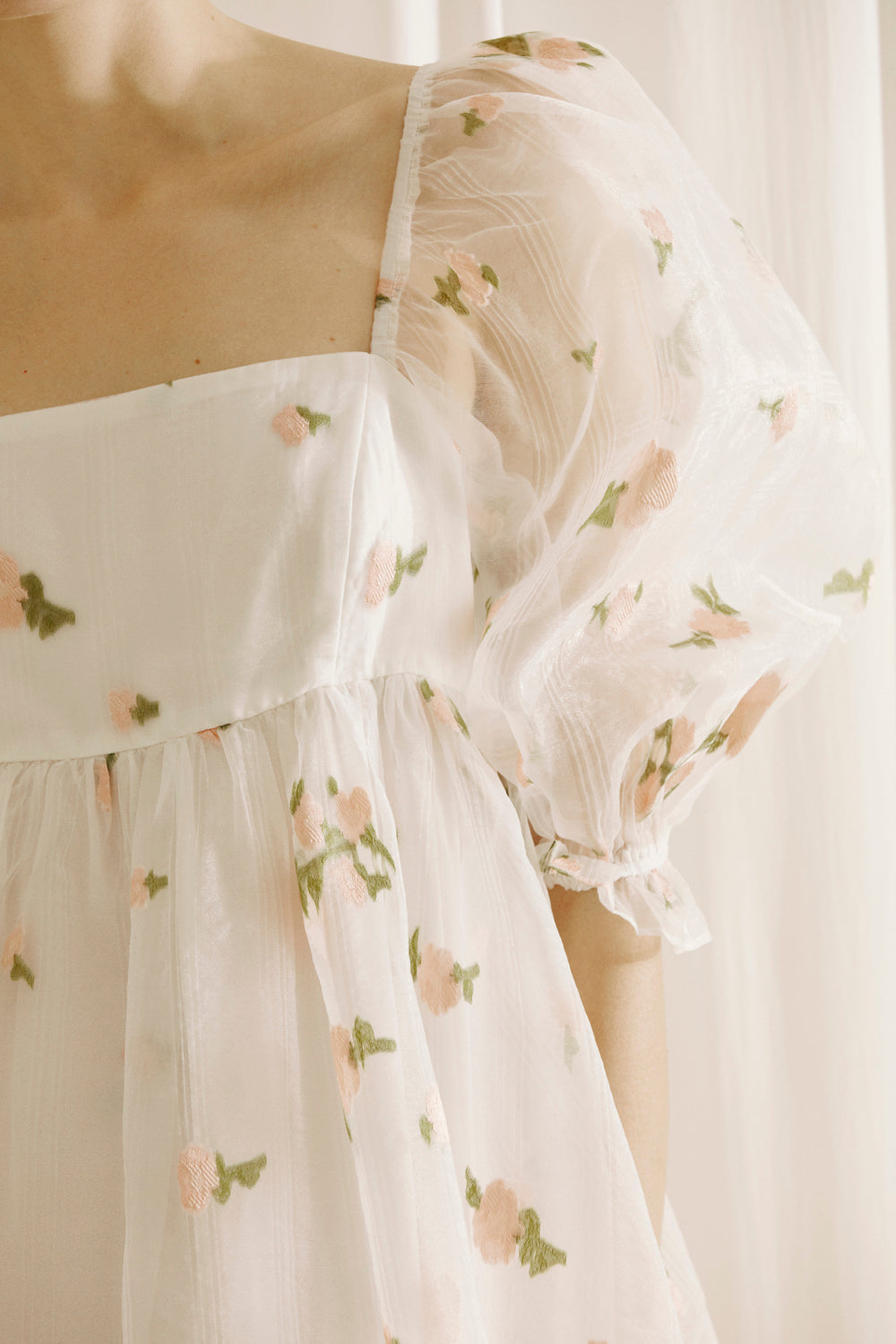 A person wearing a white dress with a top layer of a flower embroidered and textured sheer fabric, square neckline and puffy sleeves against a light curtain background