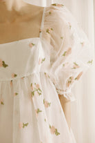 A person wearing a white dress with a top layer of a flower embroidered and textured sheer fabric, square neckline and puffy sleeves against a light curtain background