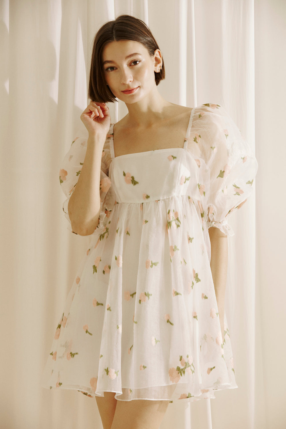 Woman wearing a white dress with a top layer of a flower embroidered and textured sheer fabric, square neckline and puffy sleeves against a light curtain background