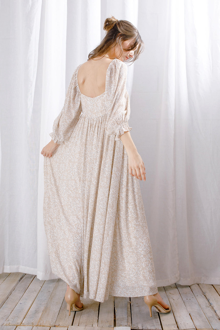 Back view of a model wearing a long, light-colored dress standing against a white curtain.