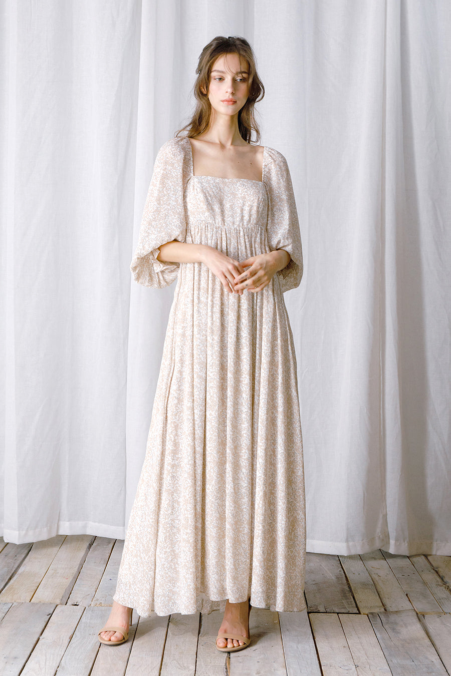 Woman wearing a long, light-colored dress standing against a white curtain.